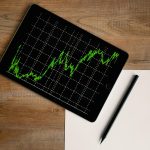 Flatlay of an iPad displaying stock market graph on a wooden desk with a pencil and paper.